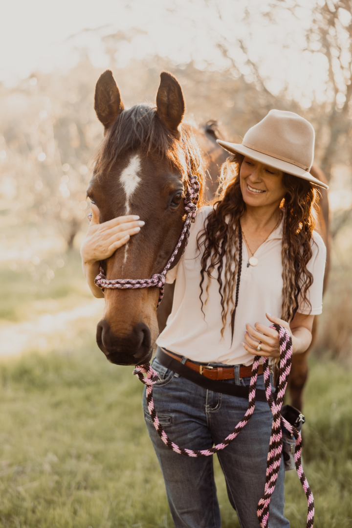 A woman smiling and gently holding a brown horse's head with a pink and black halter