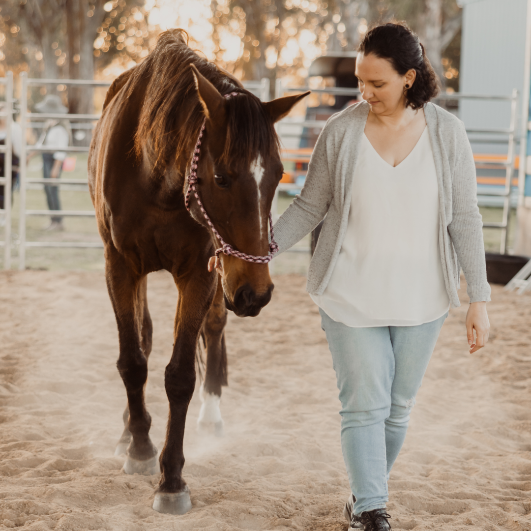 A woman with dark hair, wearing a light gray cardigan and white top, is walking alongside a brown horse with a white blaze on its face